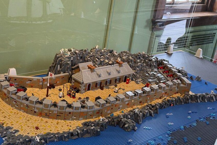 A detailed LEGO model of a coastal fortification with a central building, surrounding walls, and a sandy beach. 3