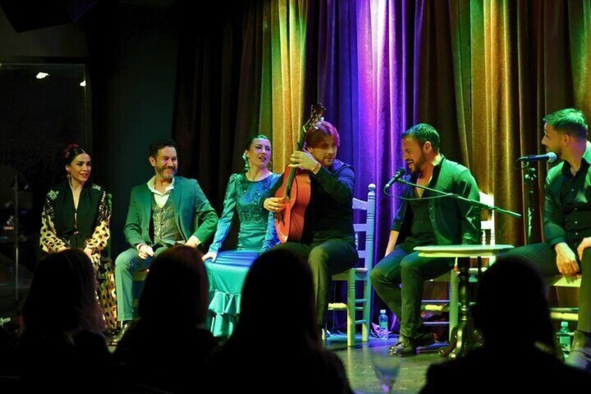 A group of people on stage with musical instruments, seated on chairs, and a microphone stand. 4