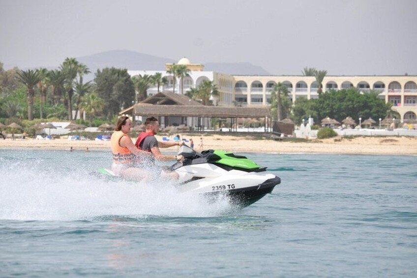 Un jet ski con dos personas, agua salpicando detrás y una playa con edificios al fondo. 3