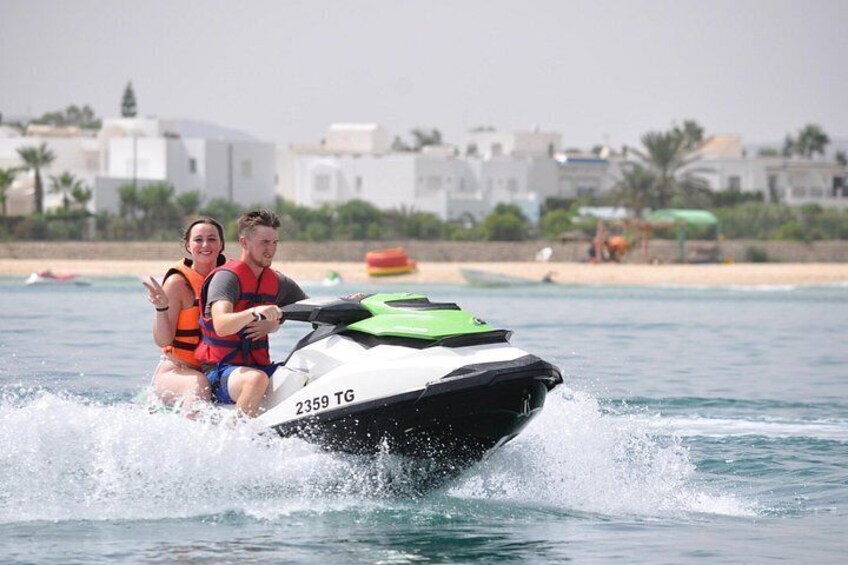 Dos personas en un jet ski, con una playa y edificios al fondo. 5