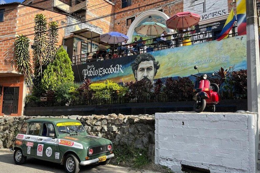 A vintage car parked in front of a mural, a scooter, and a building with a sign. 1