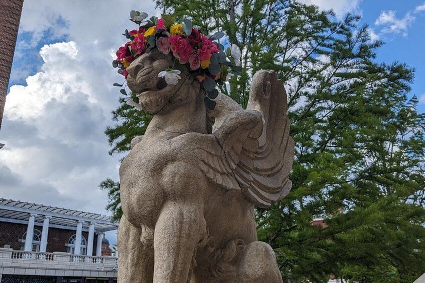 En stenskulptur som föreställer en figur med en blomsterkrans, placerad framför en bakgrund av träd och en byggnad. 4