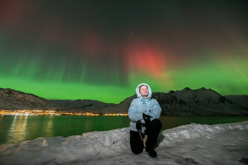 A person sitting on snow with a view of the aurora borealis over a body of water and mountains. 3