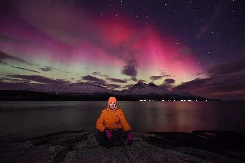 A person in an orange jacket sitting on a rock by a calm body of water with a night sky featuring the aurora borealis. 5