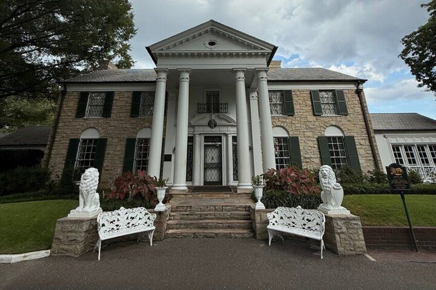 A large stone building with a central entrance, flanked by two white benches and statues. 1