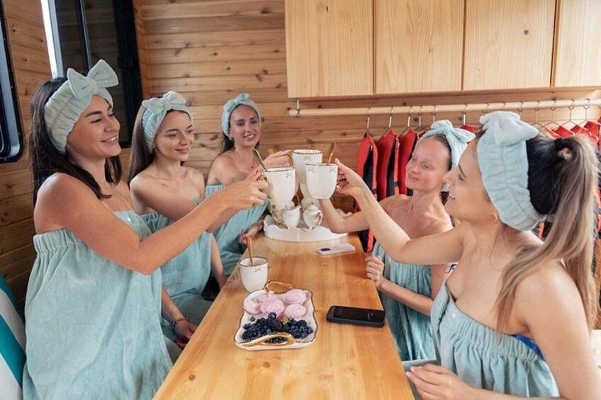 Quatre personnes, vêtues de robes bleu clair assorties et coiffées de bandeaux, trinquent avec des tasses blanches autour d’une table en bois sur laquelle est disposée une assiette de fruits. 2