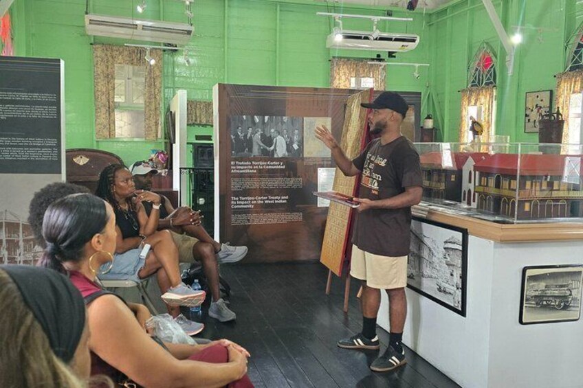 A museum exhibit with a man presenting to an audience, green walls, and historical displays. 3