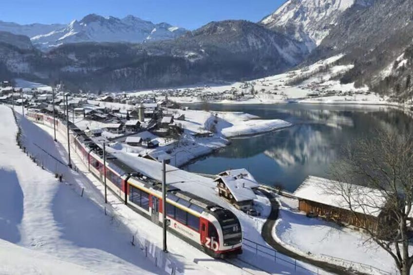Un tren sobre vías con edificios cubiertos de nieve y un lago en una zona montañosa. 4