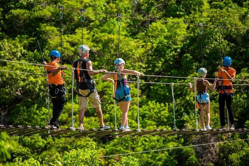 Menschen auf einem Zipline-Parcours mit Sicherheitsgurten und Helmen, umgeben von üppigem Grün. 2