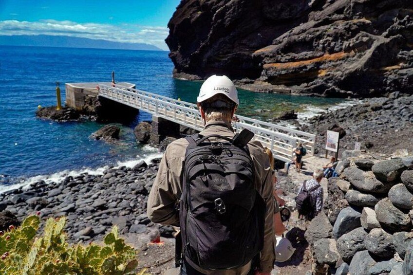 A man with a backpack and a white cap standing on a rocky beach, looking at a wooden bridge over the water. 5