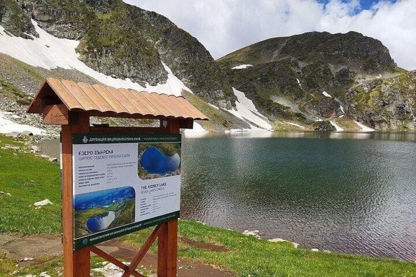 A lake with a wooden signboard in the foreground, surrounded by mountains and snow patches. 5
