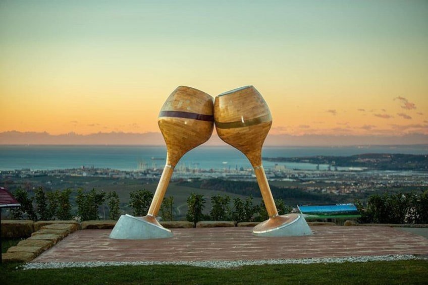 Two wooden sculptures resembling wine glasses on pedestals, set against a scenic backdrop of a cityscape at sunset. 2