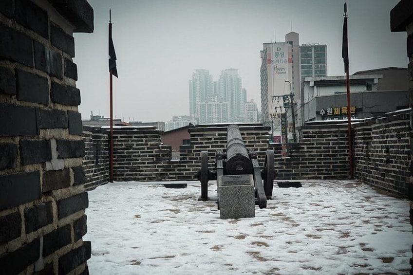 A snowy courtyard with a cannon, brick walls, and a cityscape in the background. 4