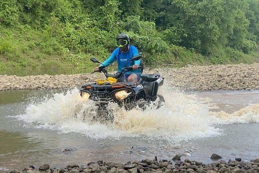 A person riding an ATV through a river, surrounded by rocks and greenery. 3