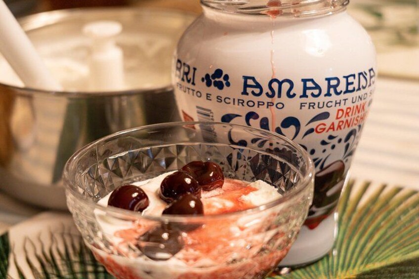 Een glazen schaal met een dessert, kersen, en een fles met de tekst 'Amarena' op de achtergrond. 3