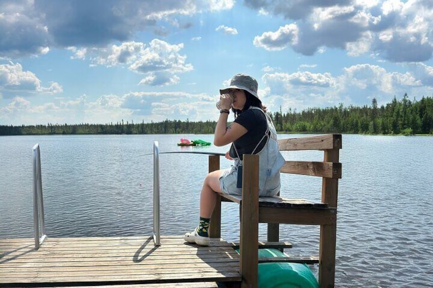 Eine Person sitzt auf einer hölzernen Mole am See, hält ein Getränk und im Hintergrund sind eine grüne Tasche und ein Paddel zu sehen. 2