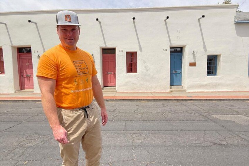 A man in an orange shirt and beige pants standing in front of a white building with red doors and windows. 1