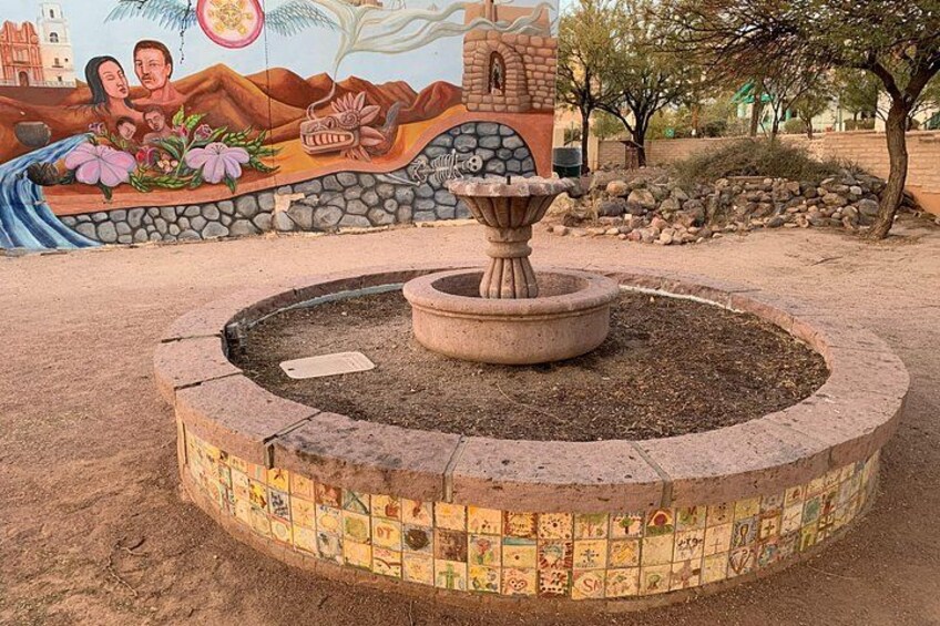A circular stone fountain with a tiled border, surrounded by a mural with cultural motifs. 5