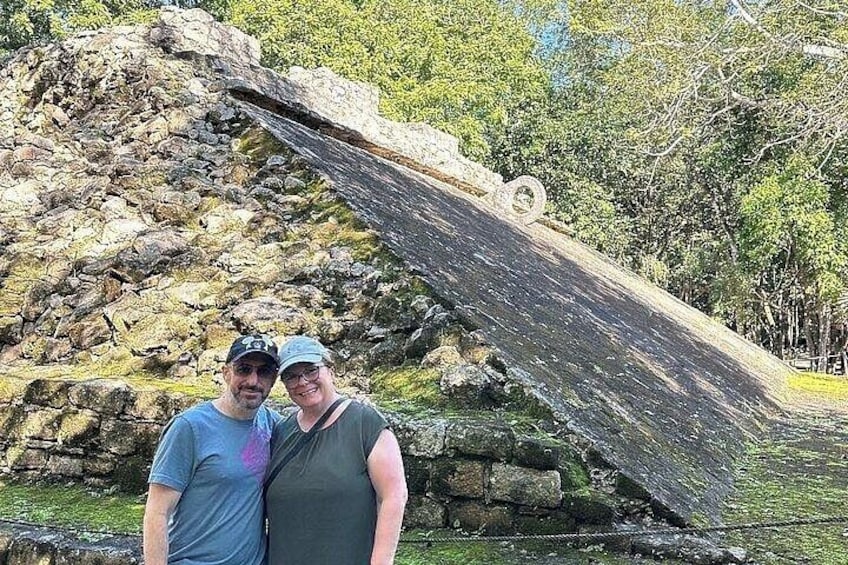 A man and a woman standing in front of a large, tilted stone structure with moss and greenery around it. 5