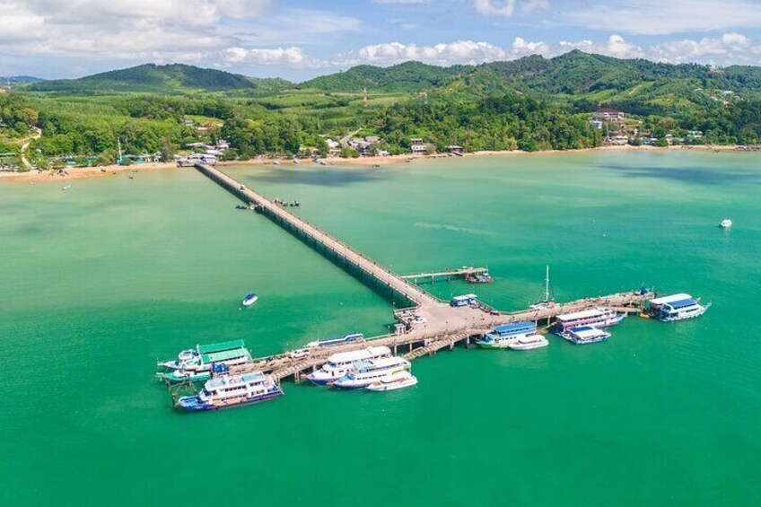 An aerial view of a coastal area with a pier extending into turquoise waters, surrounded by boats and lush green hills in the background. 2