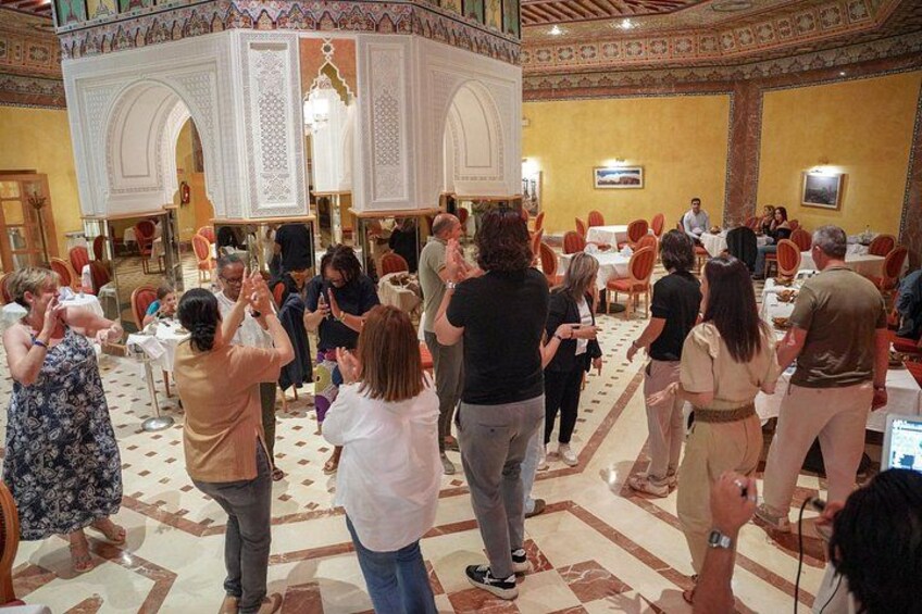 A group of people in a large hall with a patterned floor, tables, and chairs. 2