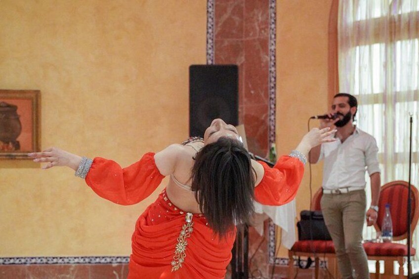 A person in a red dress with gold embroidery, a man in a white shirt, and a microphone stand in a room with a marble floor and a painting on the wall. 4