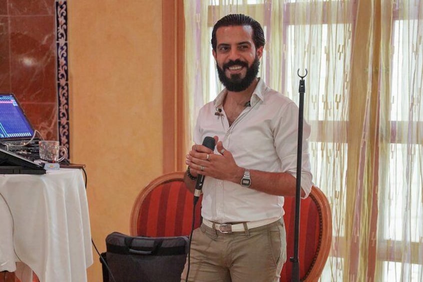 A man holding a microphone, standing in a room with a laptop, a microphone stand, and a table with a white cloth. 2