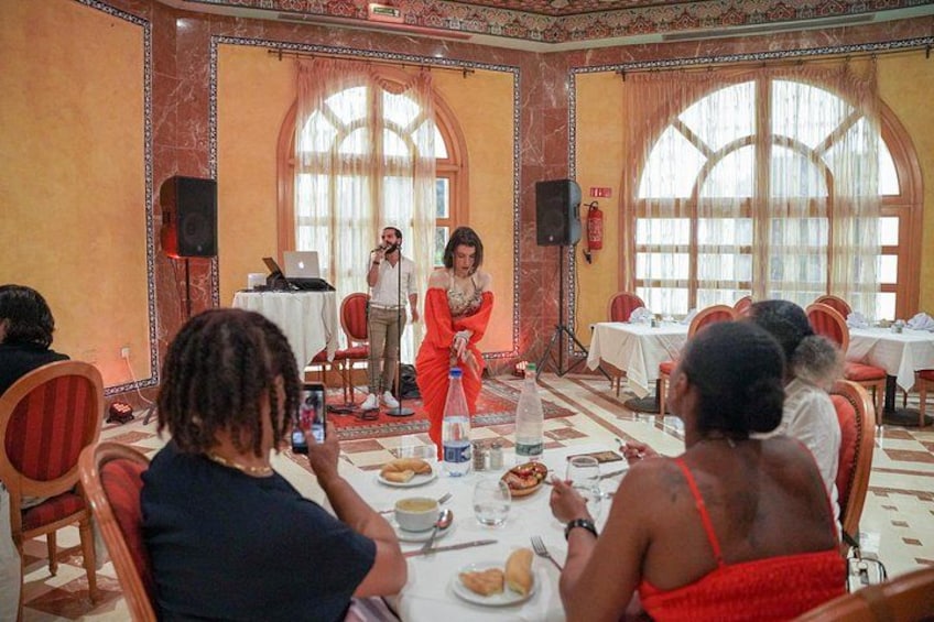 A woman in a red dress speaking at a microphone, with an audience seated at tables in a room with ornate walls and large windows. 4