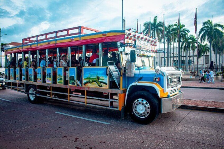 A colorful bus with passengers, parked on a street with palm trees and buildings in the background. 1