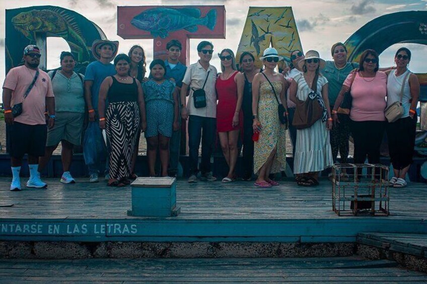 A group of people standing on a wooden deck with a sign that reads "ENTARSE EN LAS LETRAS" and a large letter 'A' in the background. 5