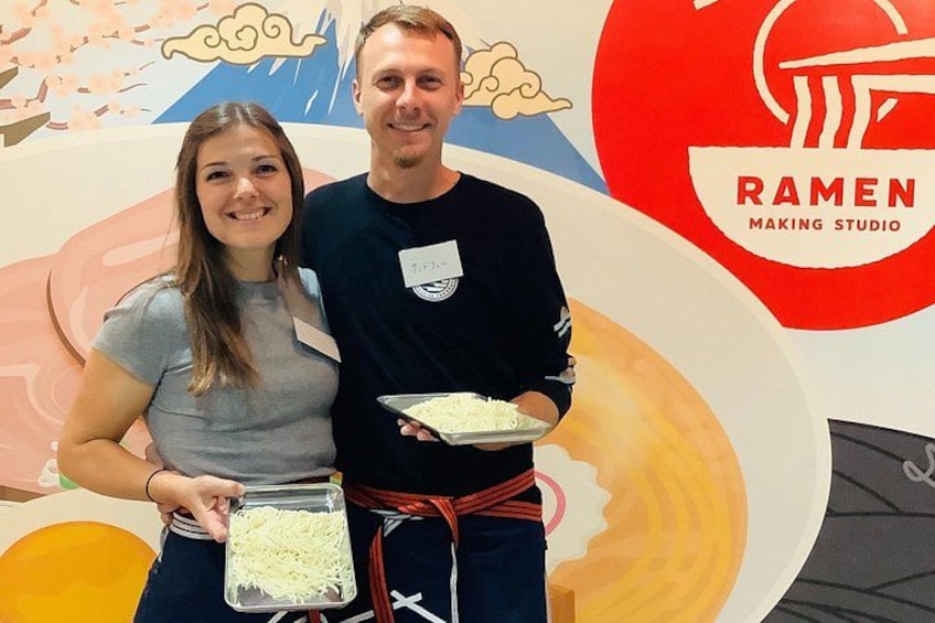 Dos personas sosteniendo bandejas con comida frente a un letrero de restaurante de ramen. 2