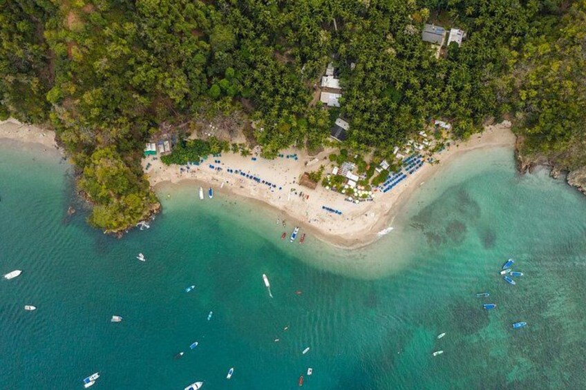An aerial view of a beach with clear turquoise waters, a sandy shore, and a small cluster of buildings surrounded by dense greenery. 3