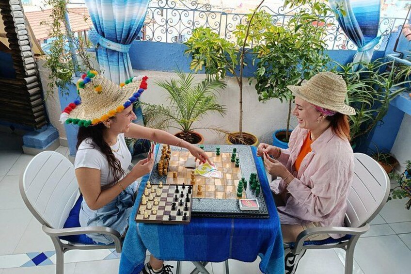 Zwei Personen spielen Schach an einem Tisch im Freien mit einer blauen Tischdecke, umgeben von Blumentöpfen und einem blauen Geländer. 3