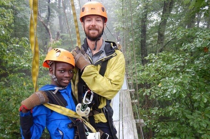 Two people in a forest, wearing helmets and harnesses, on a suspended bridge. 3