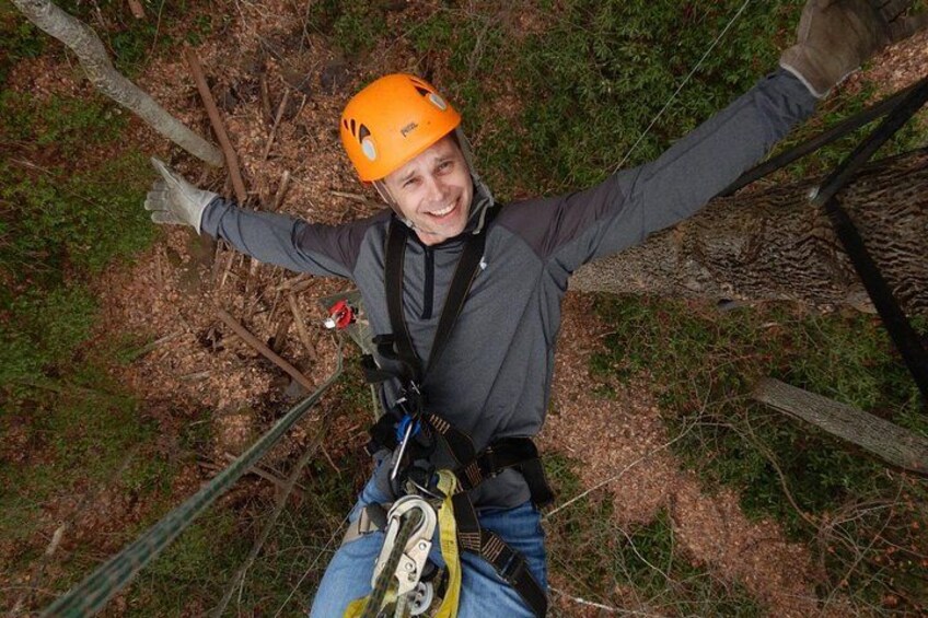 A person bungee jumping with arms outstretched, wearing an orange helmet and harness, with a forested background. 4