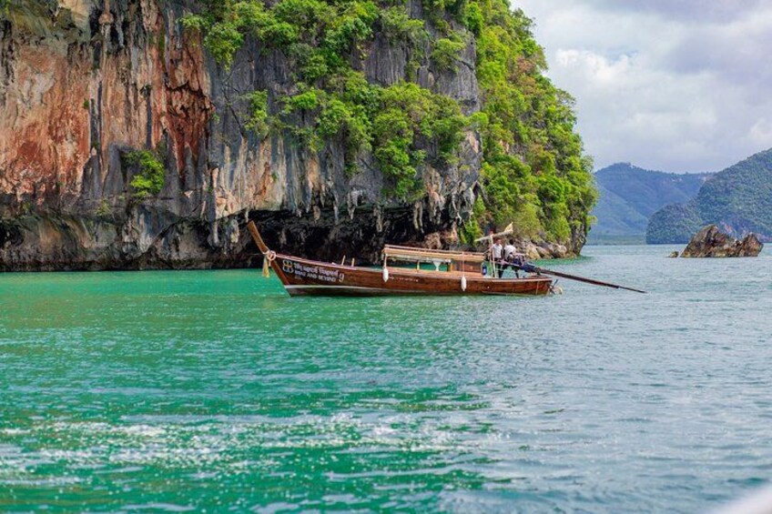 Un bateau en bois traditionnel sur une eau turquoise, une falaise rocheuse parsemée de verdure et des montagnes lointaines. 2