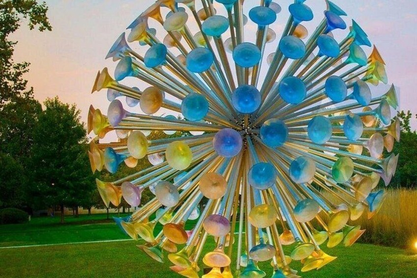 A large, colorful spherical sculpture with a central chain, surrounded by a grassy field and trees. 3