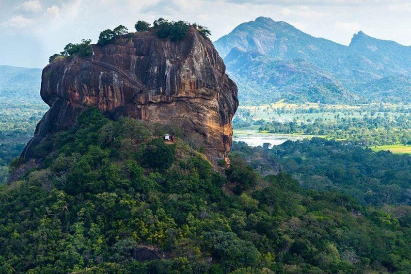 A large rock formation with a small building on top, surrounded by dense forest and mountains in the background. 1