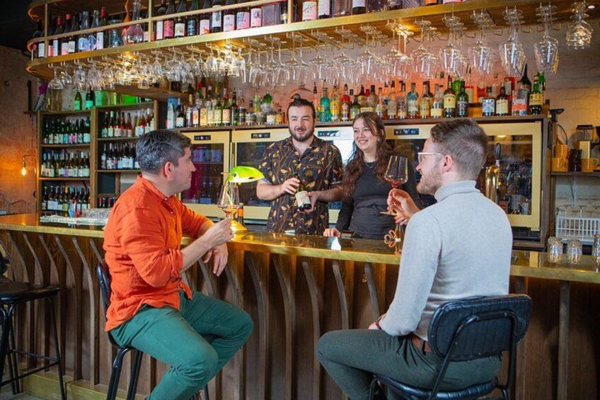 Un bar où les clients sont assis et debout, tiennent des verres à la main, et un barman se tient derrière le comptoir. 3