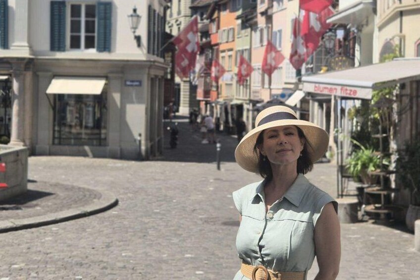Eine Frau mit Hut steht auf einer Kopfsteinpflasterstraße, im Hintergrund sind Gebäude und Flaggen zu sehen. 2