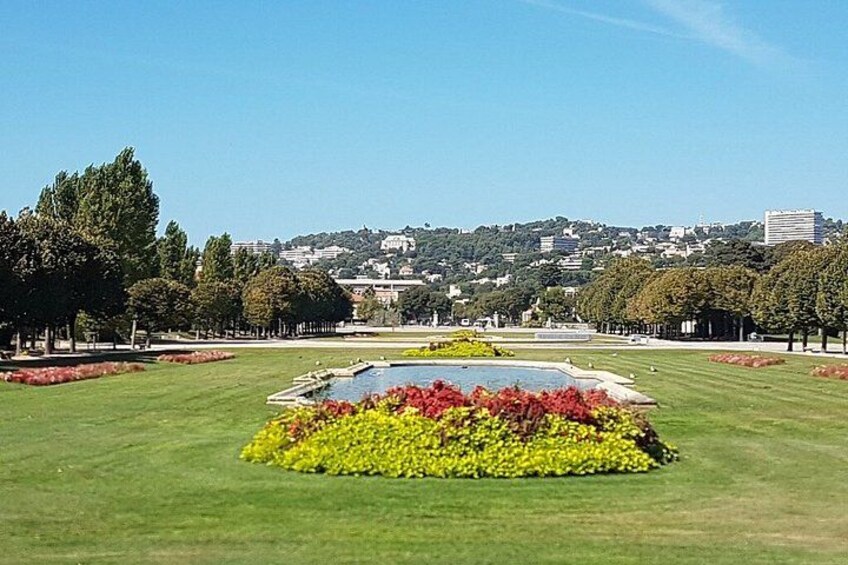 Ein Park mit einem zentralen Blumenbeet, grünen Rasenflächen und einer in der Ferne liegenden Stadtansicht. 2