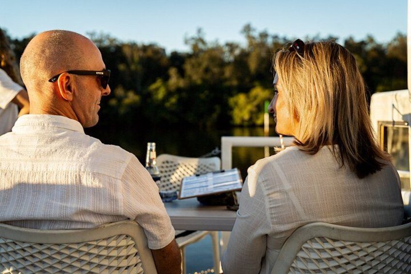 Two people sitting at an outdoor table with a laptop and papers, surrounded by greenery. 4
