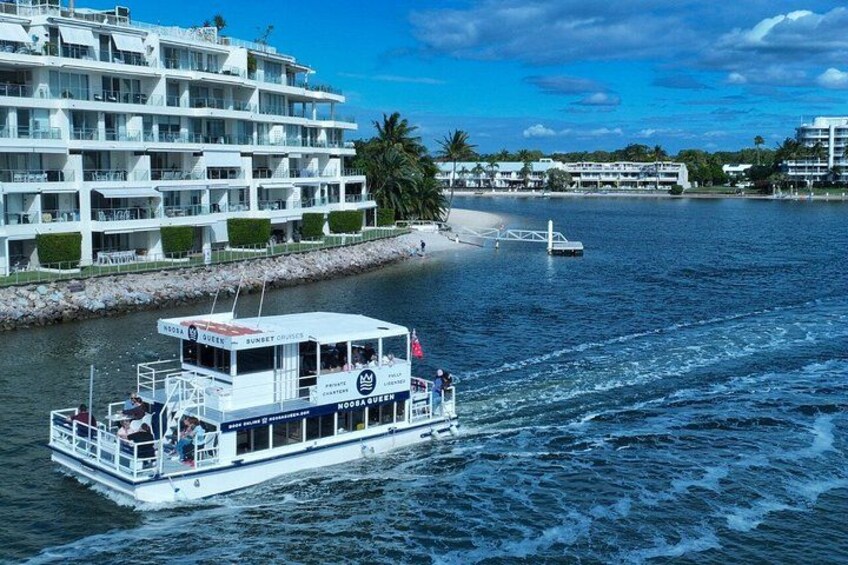 A white boat with 'MOORA QUEEN' written on it, moving on a body of water near a waterfront building and a beach. 1