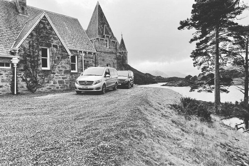 A stone building with a steep roof, parked cars, and a body of water in the background. 2