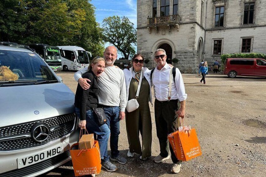 Four people standing in front of a silver Mercedes-Benz with a license plate V300 MAC, holding orange shopping bags. 2