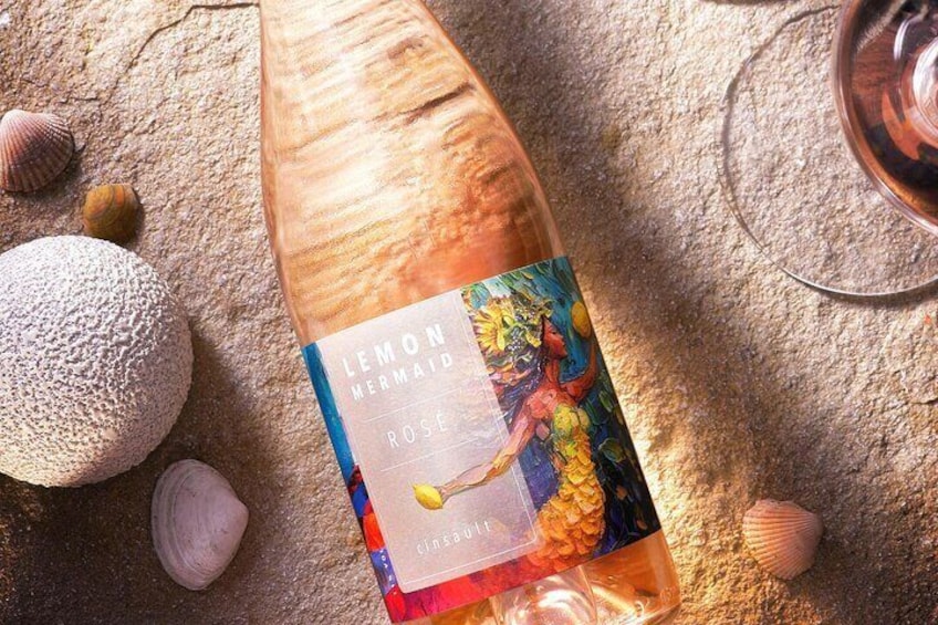 Una botella de vino 'Lemon Mermaid Rose' con una etiqueta colorida, sobre una superficie texturizada rodeada de conchas marinas. 3