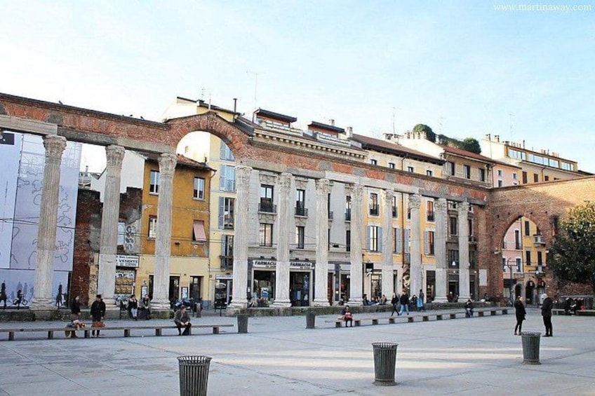 Una piazza storica caratterizzata da architettura classica, con colonne e archi, e persone sedute o in movimento. 3