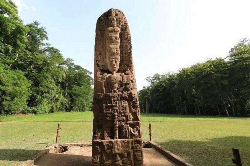 A tall stone monument with intricate carvings, surrounded by a grassy area with trees in the background. 3