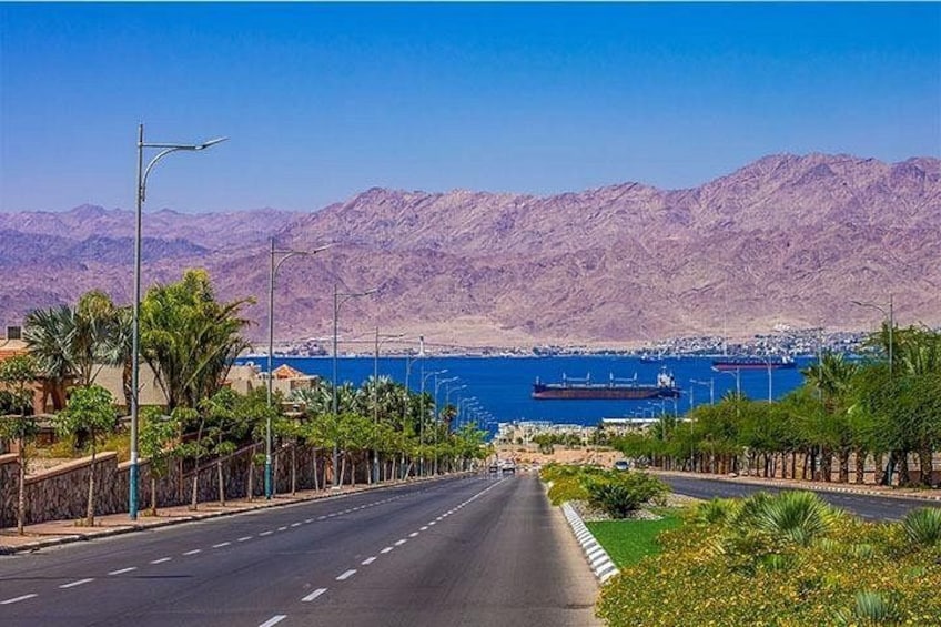 A coastal road with mountains in the background, a large body of water, and a cargo ship. 2