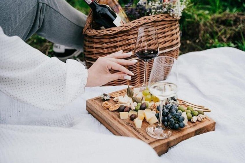 En picknickuppsättning med en träbricka som innehåller ost, vindruvor och nötter, en korg i bakgrunden och en flaska vin. 1